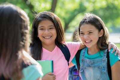 Two smiling students with backpacks outdoors, one holding a notebook, with a third person blurred in the foreground