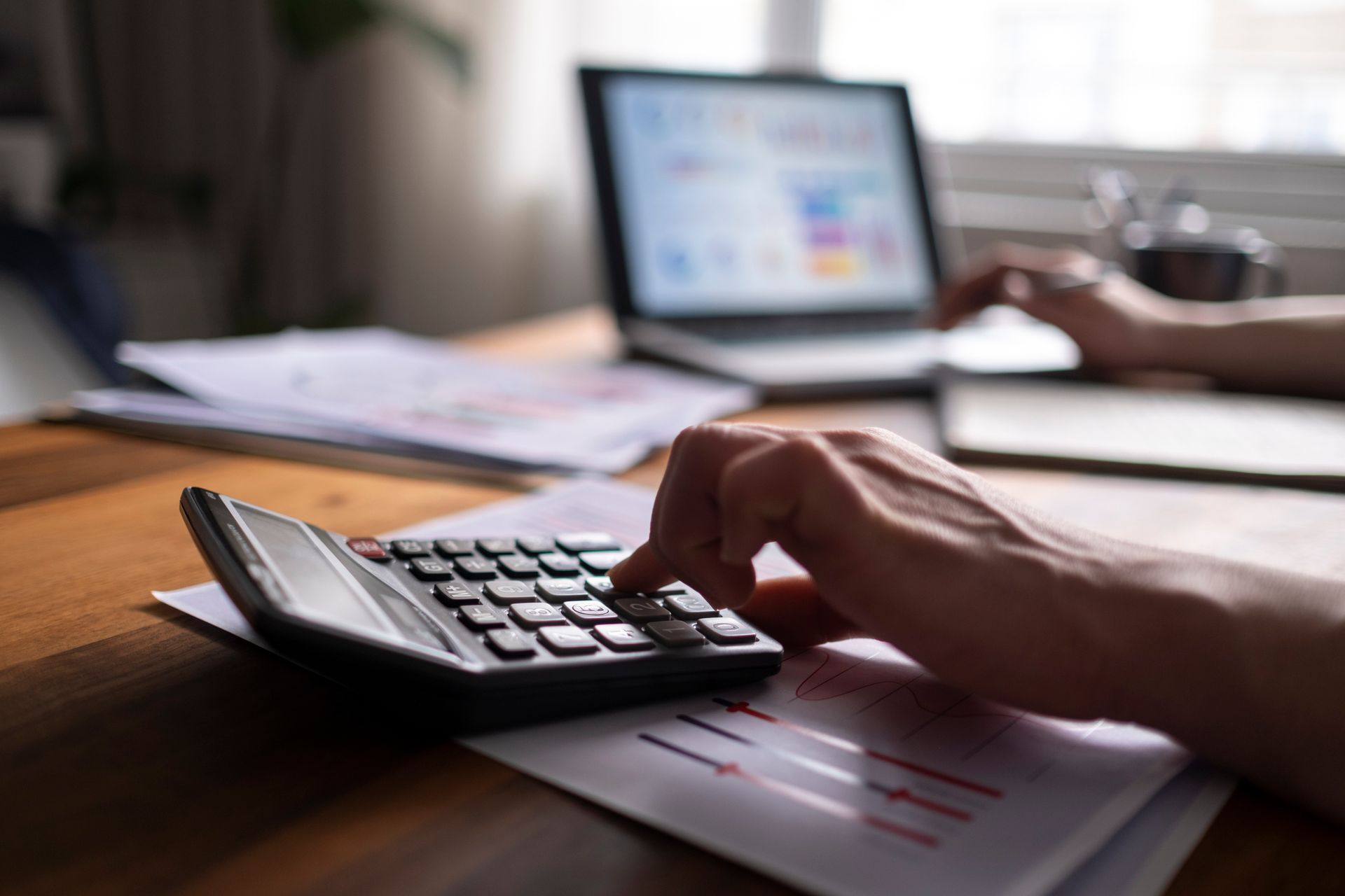 A person is using a calculator at a desk in front of a laptop.