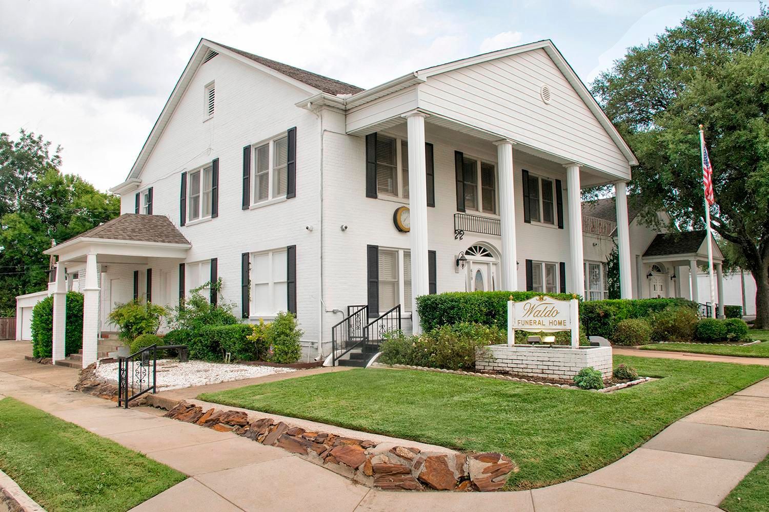 White two-story building with columns and black shutters; grassy lawn, sign reads