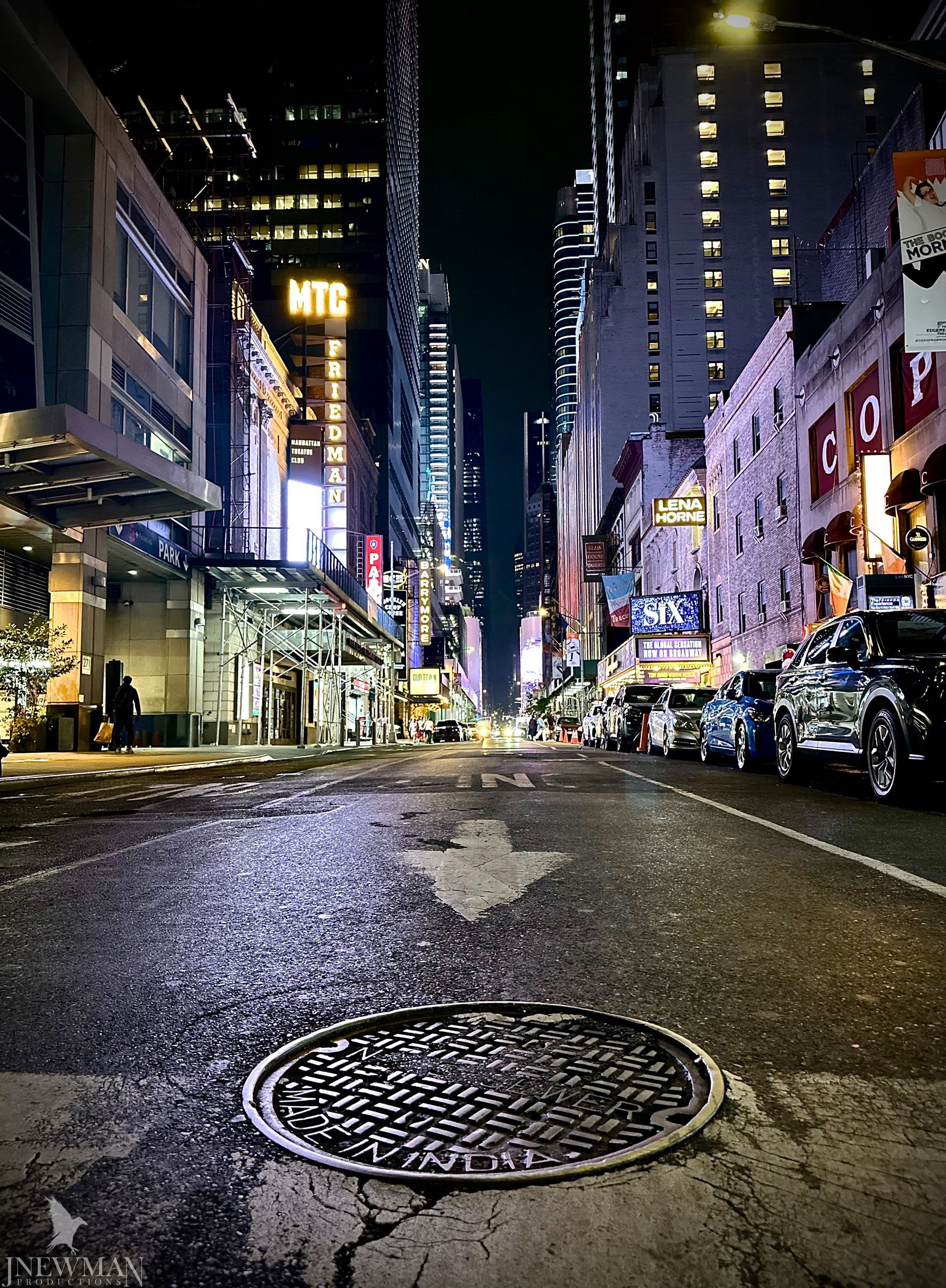 Night view down a wet city street towards brightly lit skyscrapers. Arrow on the road points to a manhole cover.