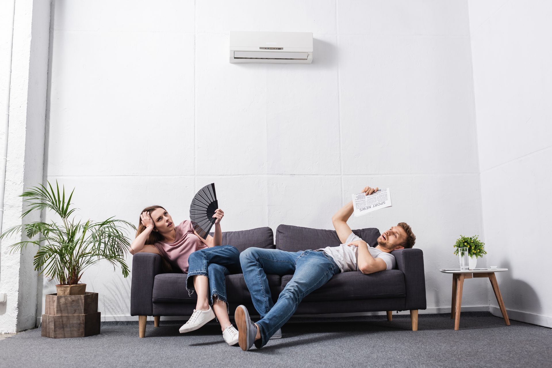 Young couple with hand fan and newspaper suffering from heat at home with broken air conditioner.