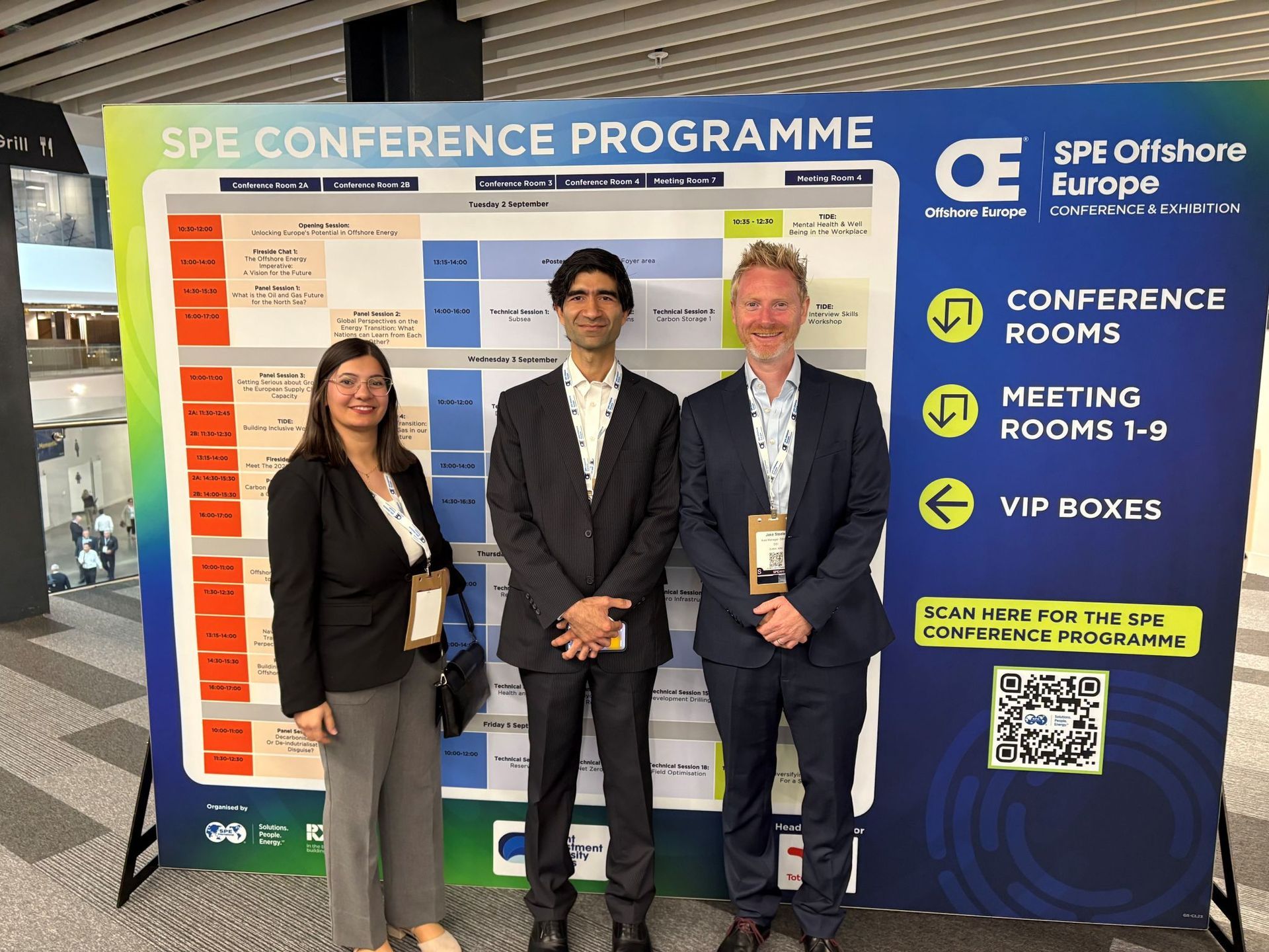 Three people stand in front of a conference schedule board at the SPE Offshore Europe event.