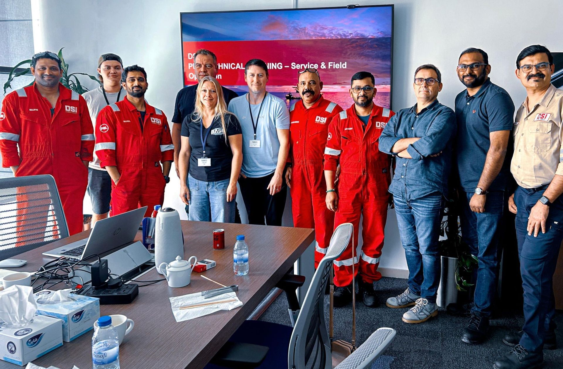 Group of people, some in red jumpsuits, posing in an office setting with a large screen displaying text and a nautical image.