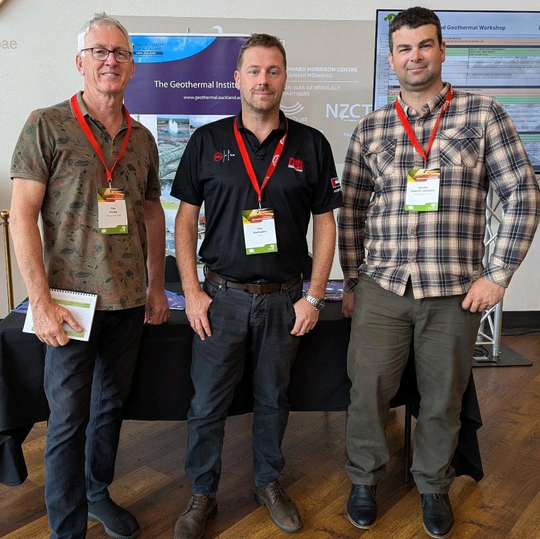 Three men stand by a table at a conference, wearing lanyards. The background includes a banner and screen.