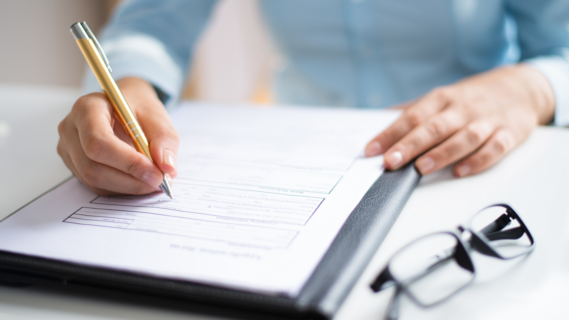 Person writing on a document with a pen, glasses are in the bottom right.