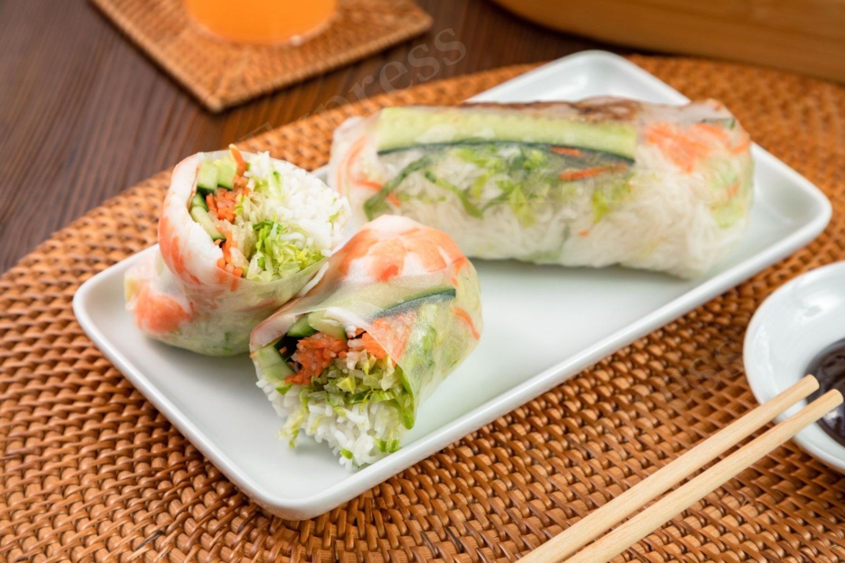 Three Vietnamese fresh spring rolls with shrimp and vegetables on a white plate, set on a woven mat with chopsticks.