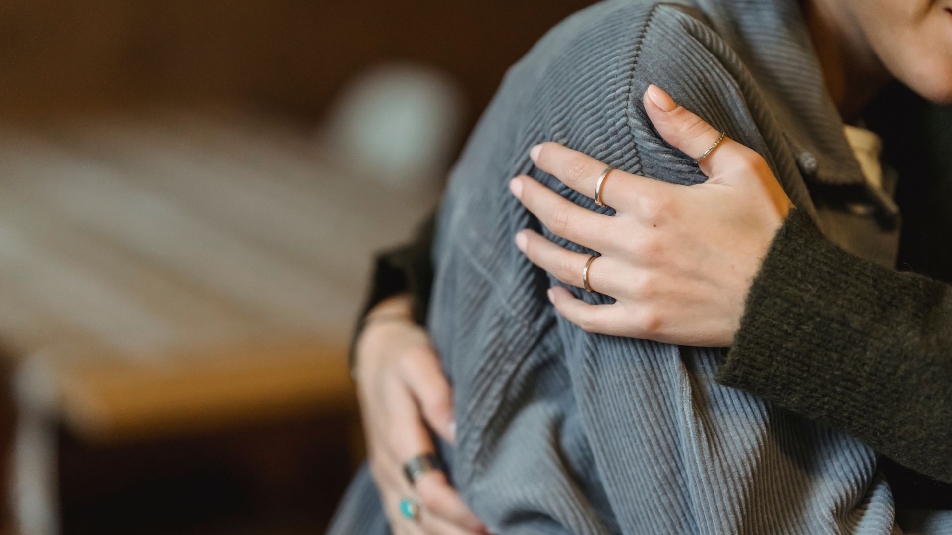 Person's hand with rings on shoulder of someone wearing a gray jacket, offering comfort.