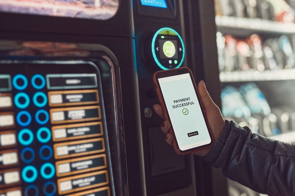Person using smartphone to pay at a vending machine with a payment gateway app open.