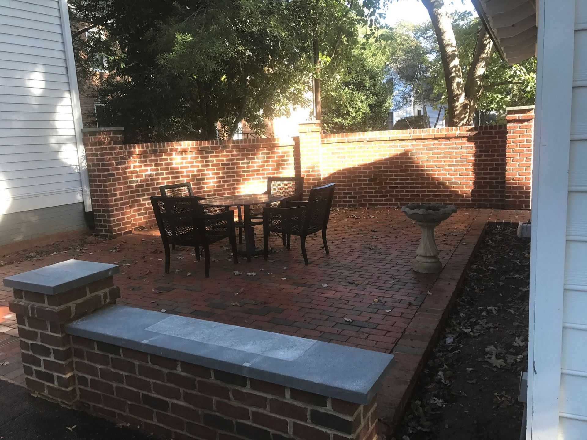 Brick patio with black chairs and table, enclosed by a low brick wall and trees casting shade