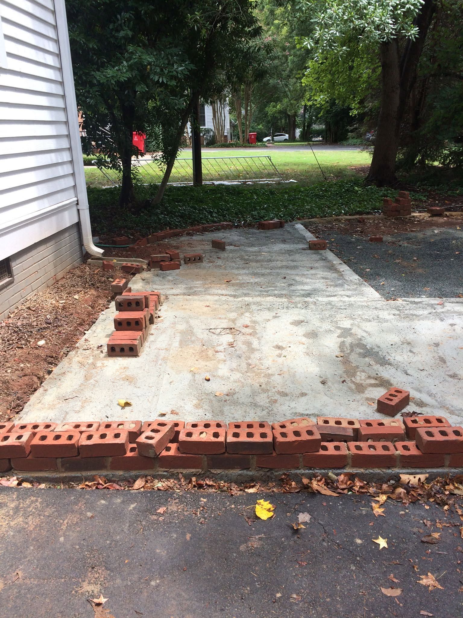 Partially built brick border around a concrete driveway beside a house and yard, with loose bricks scattered around