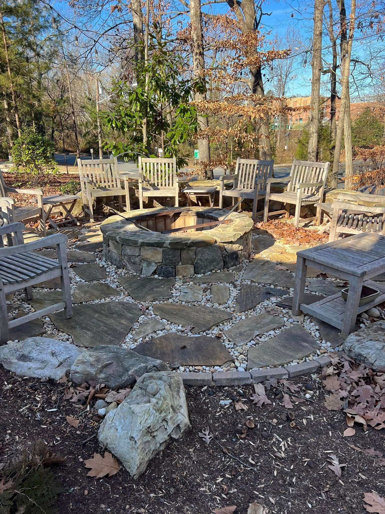 Stone patio fire pit surrounded by wooden chairs in a wooded backyard, with fallen leaves and trees.