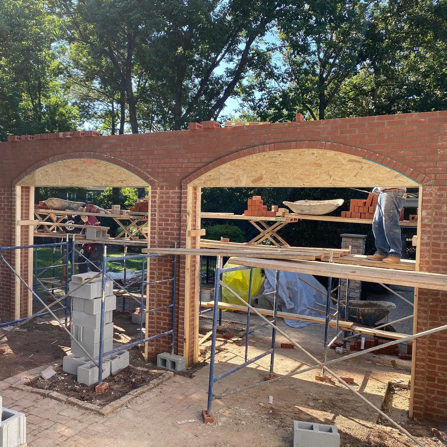 Brick building under construction with two arched openings, scaffolding, and workers inside a wooded outdoor site