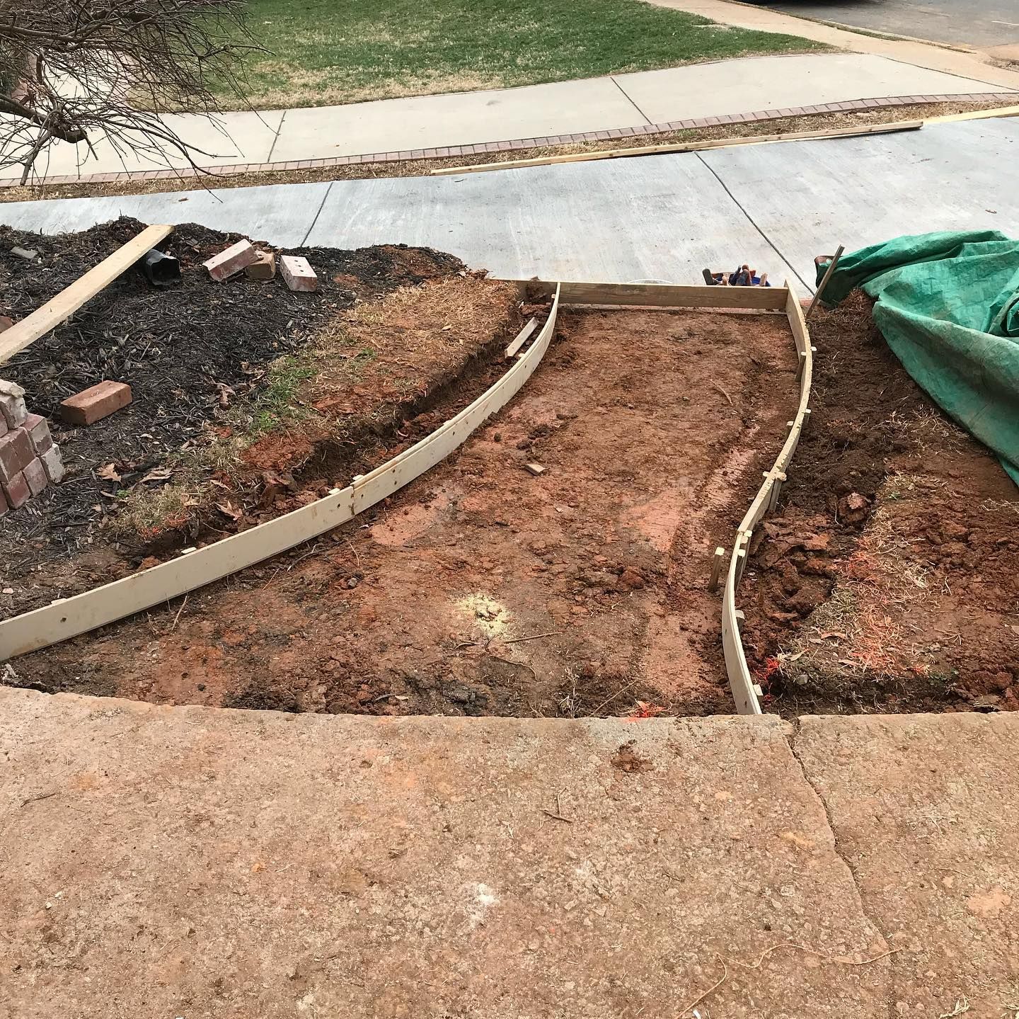 Partially built concrete walkway with dirt-filled section, edging forms, and a green tarp beside a curb