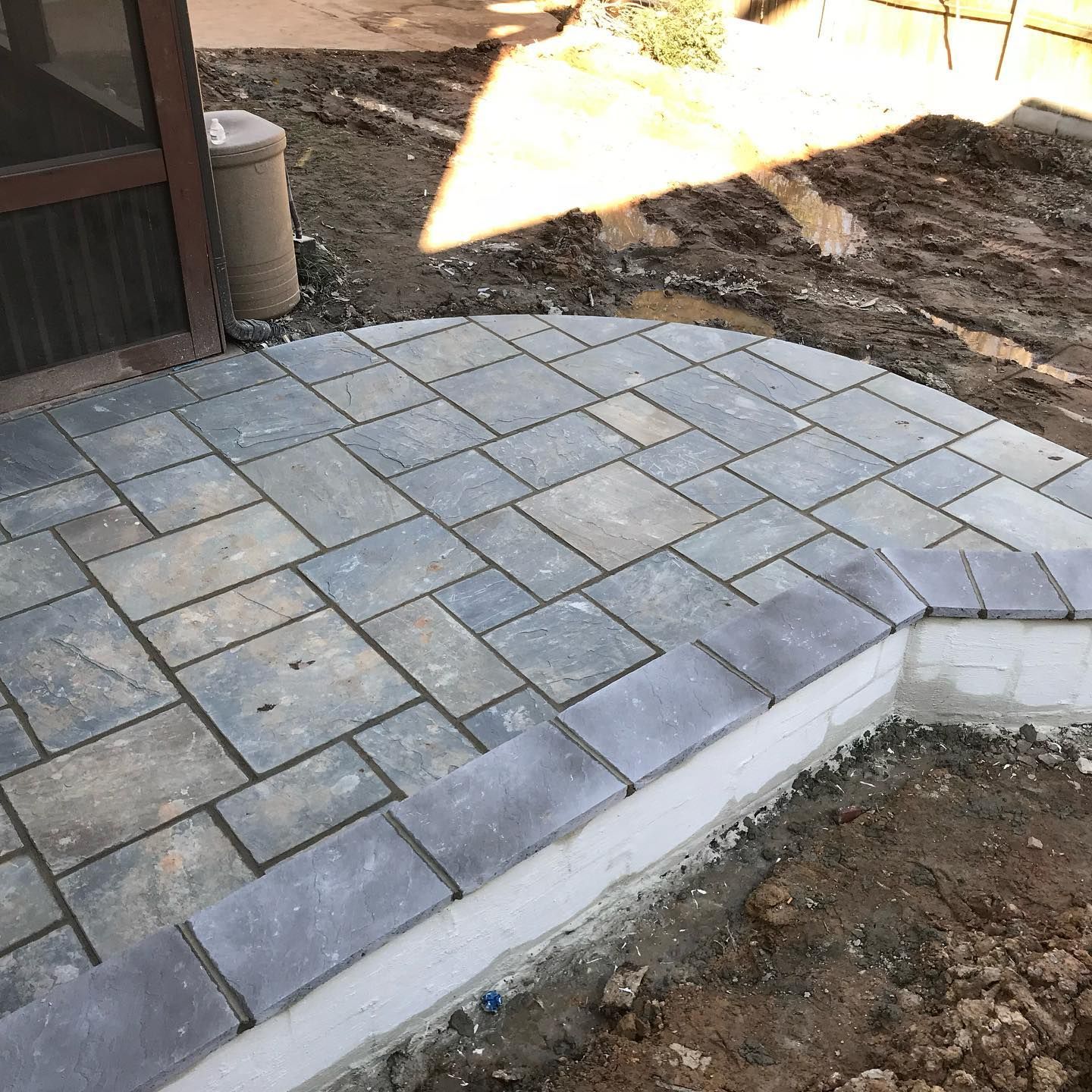 Stone paver patio with a curved edge beside a dirt yard and a building corner