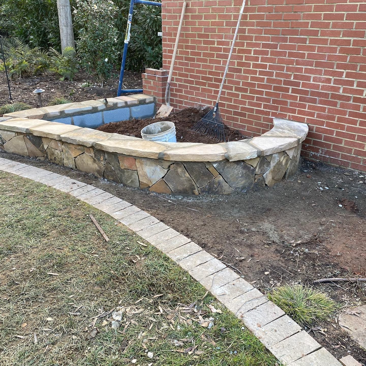 Raised stone garden bed beside a brick wall, with mulch, a small planter, and a curved paver border