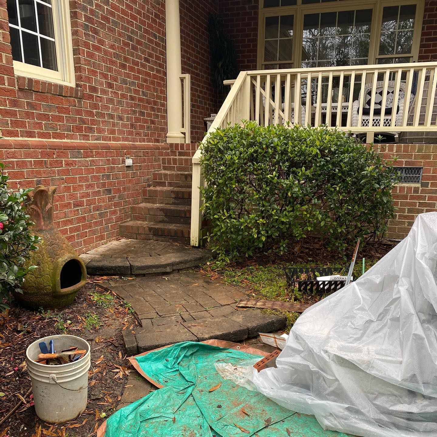 Front porch with brick wall, white railing, bushes, buckets, and tarp-covered items on the ground