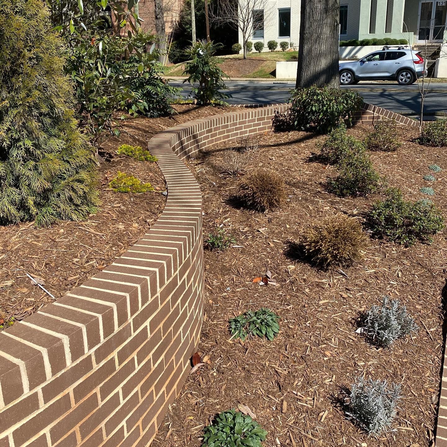 Curved brick retaining wall beside a mulched garden bed with shrubs and a parked car in the background