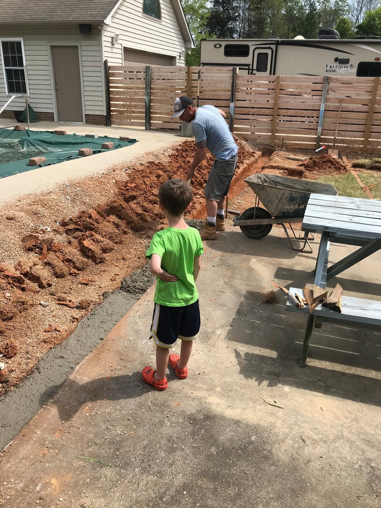 Two people shoveling dirt beside a pool and wooden fence, with a child watching nearby.