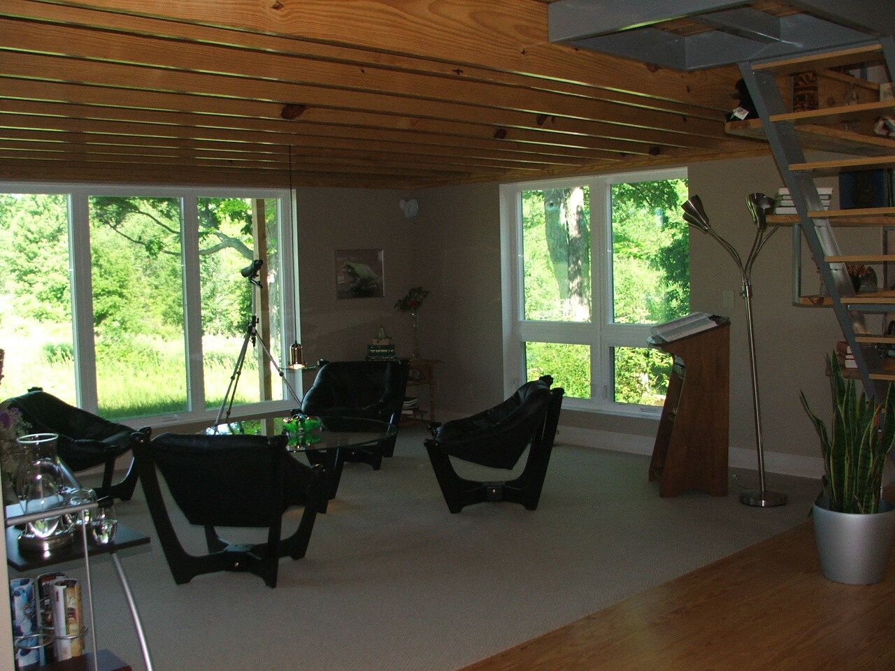 Spacious living room with large windows, black chairs, and a wood-paneled ceiling overlooking greenery.