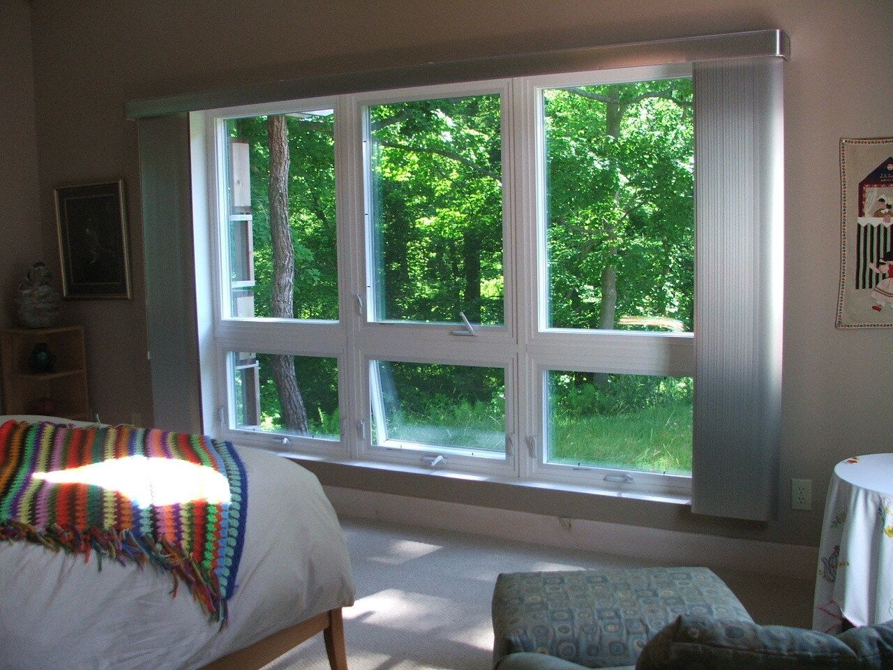 Bedroom with a large window, sheer curtains, and sunlight shining on a bed and chair.