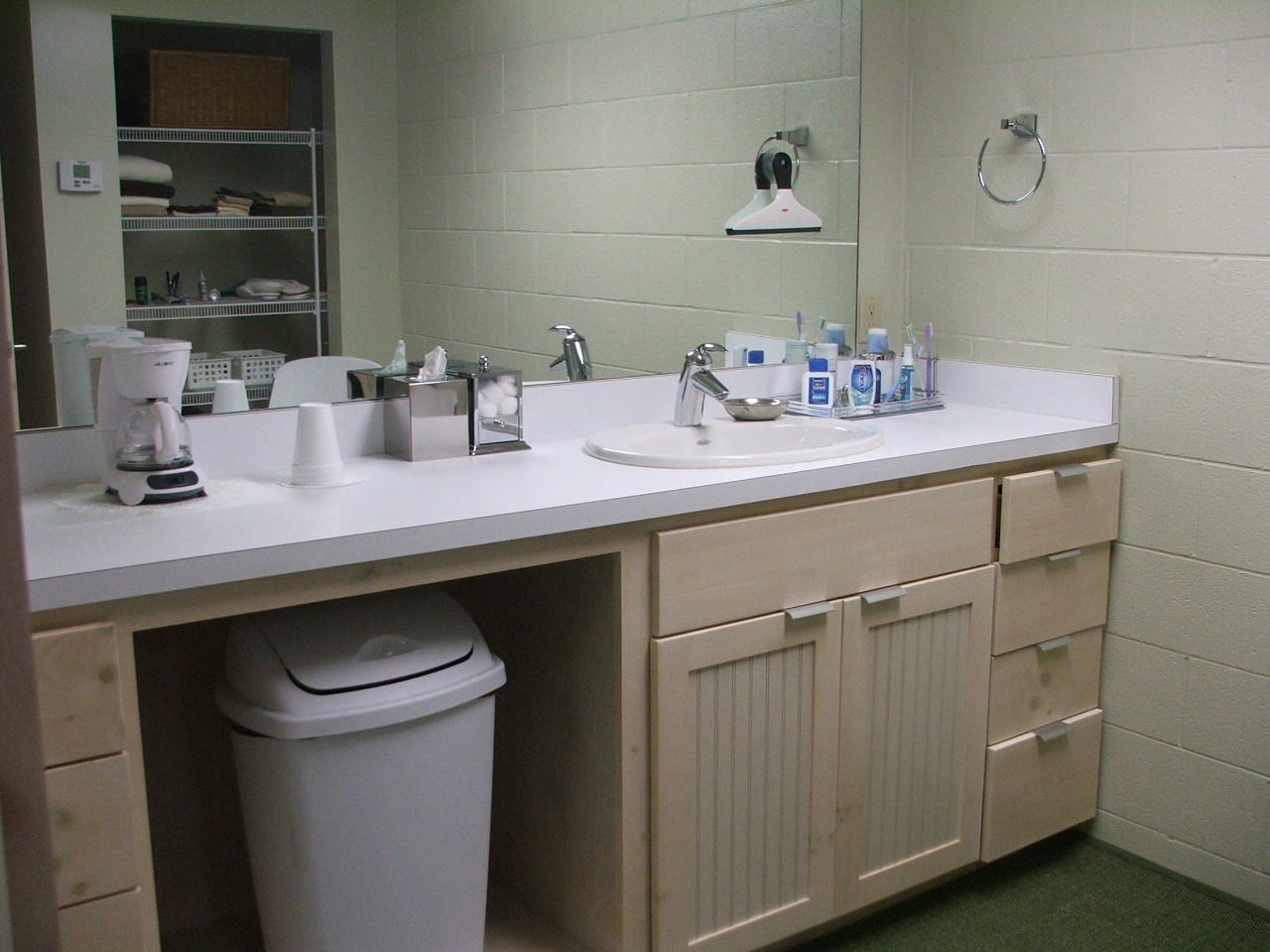 Public restroom sink area with mirror, counter, soap bottles, and trash can beneath.