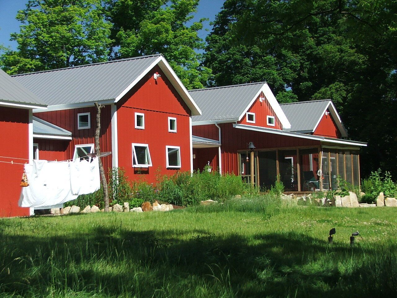 Red houses with white trim and porches in a green yard under trees and a blue sky
