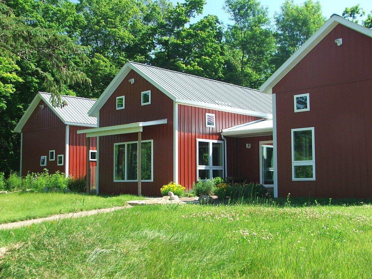Red house with white trim in a grassy yard, surrounded by green trees under bright sunlight