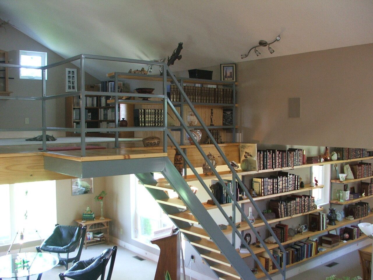 Lofted library with bookshelves, a metal staircase, and large windows in a bright open room