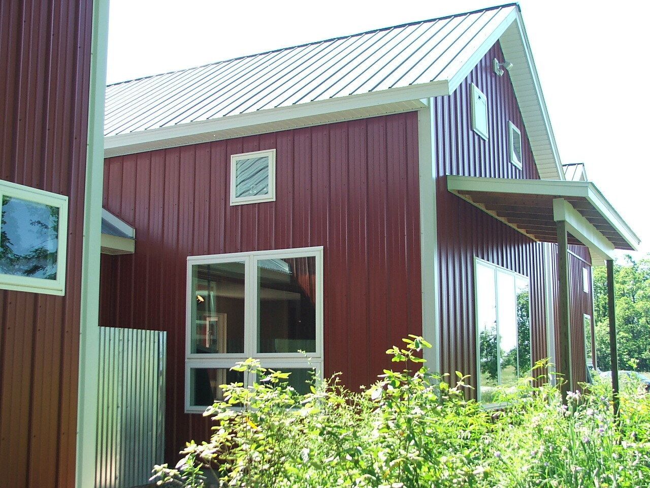 Red barn-style house with white trim and large windows, partly hidden by green bushes under a bright roof.