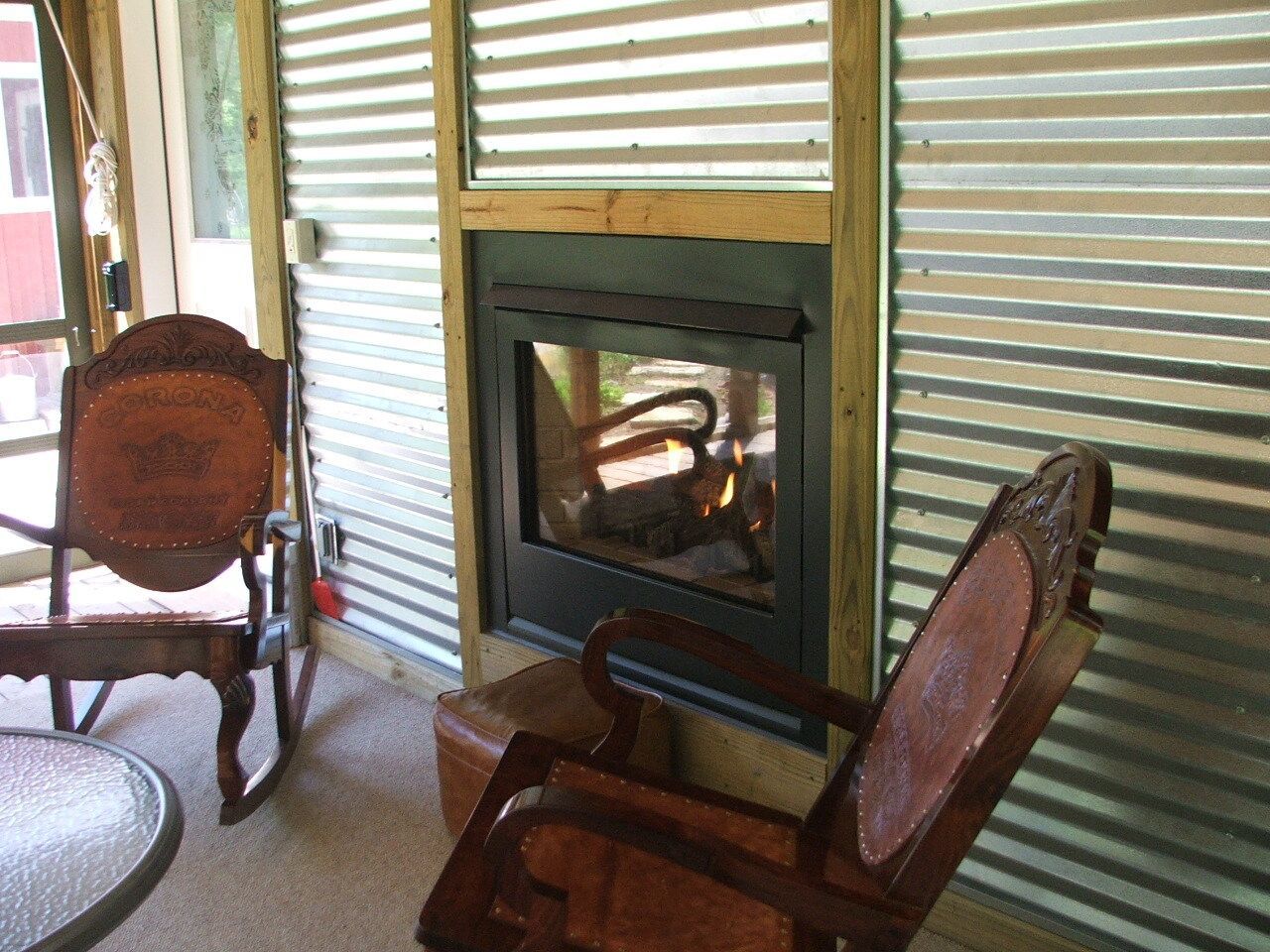 Two wooden chairs facing a small glass-front fireplace in a cozy room with corrugated metal walls