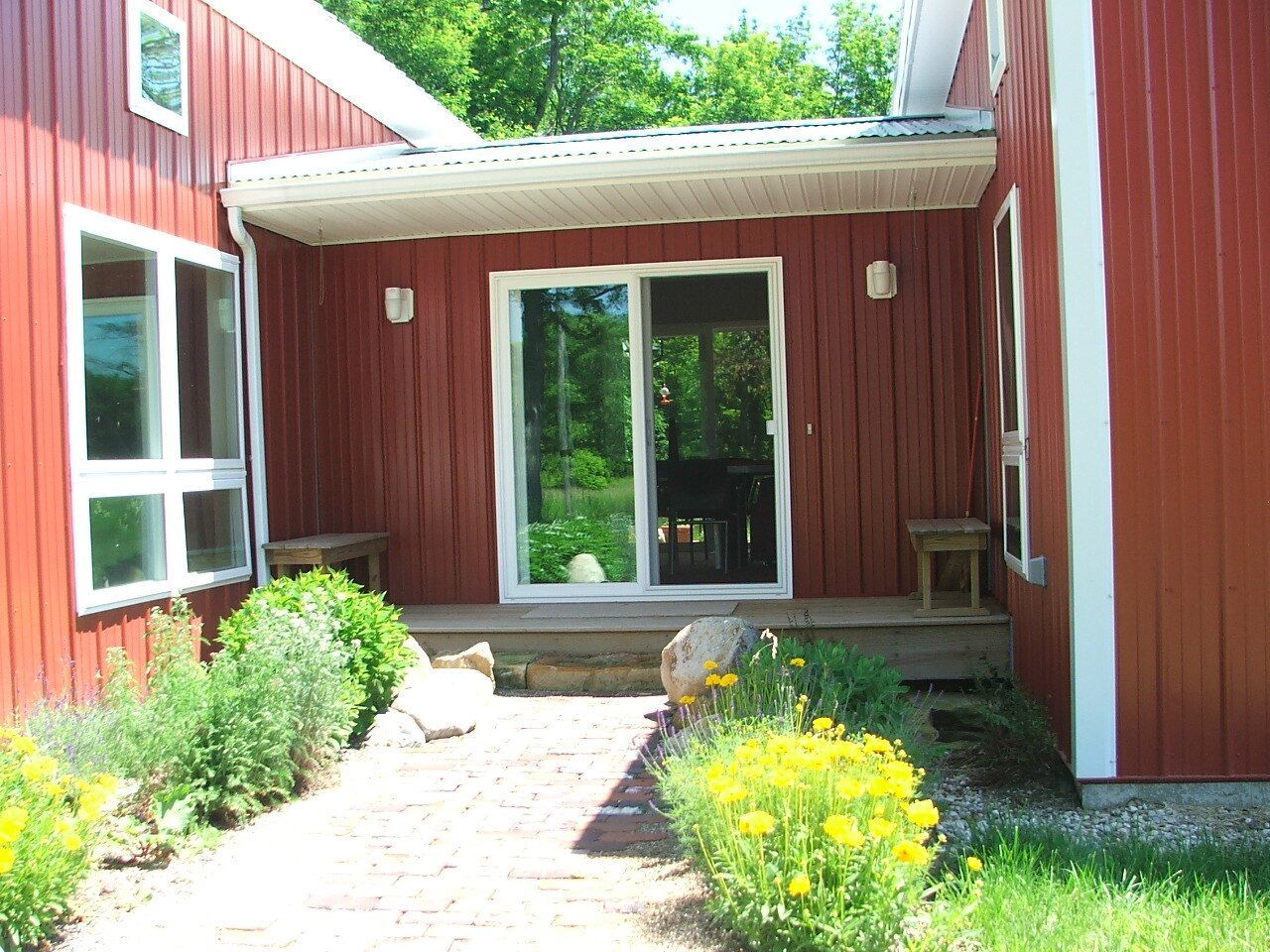 Red-sided building entrance with concrete porch, glass doors, and flower-lined walkway