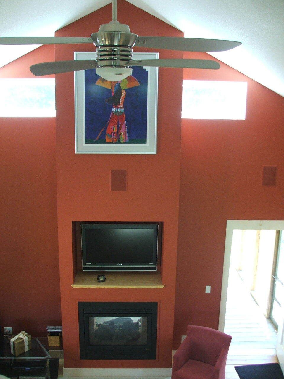 Living room with orange walls, fireplace and TV, ceiling fan, red chair, and bright windows.