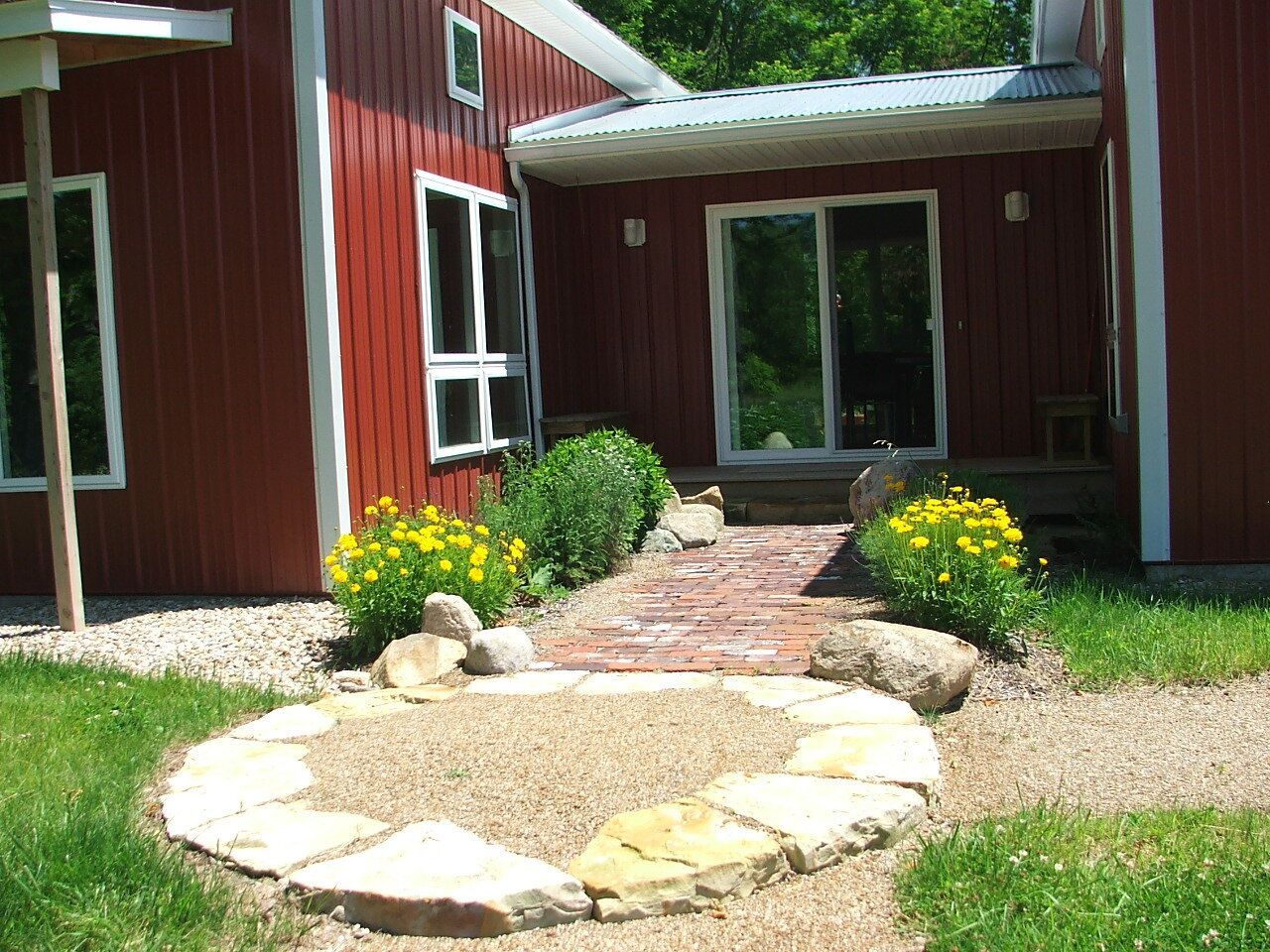 Red house with a stone walkway leading to sliding glass doors and flower beds in a yard