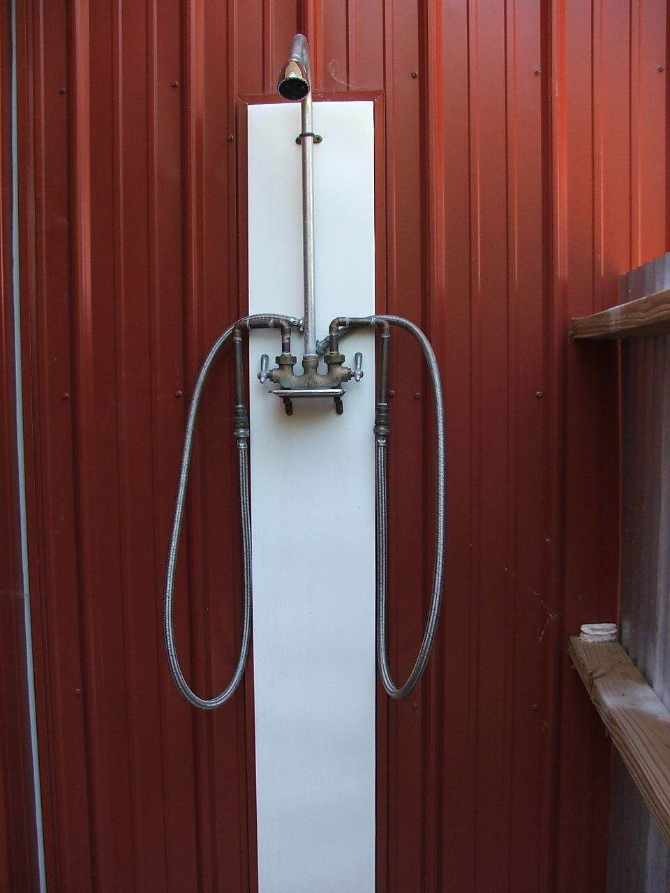 Wall-mounted outdoor shower with white panel and silver hoses against a red wooden wall.