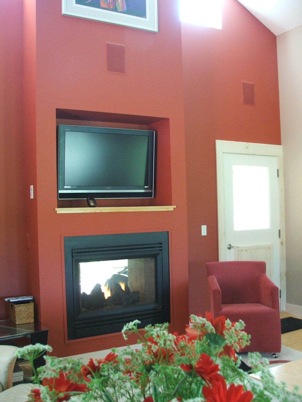Living room with red walls, wall-mounted TV above fireplace, and red armchair near a bright door.