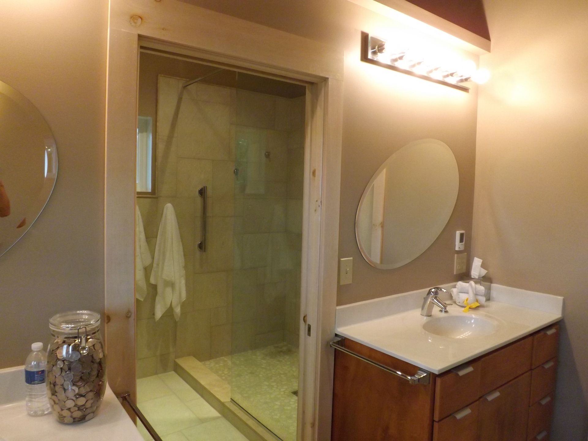 Bathroom vanity with oval mirror and lit wall sconce beside a glass shower door
