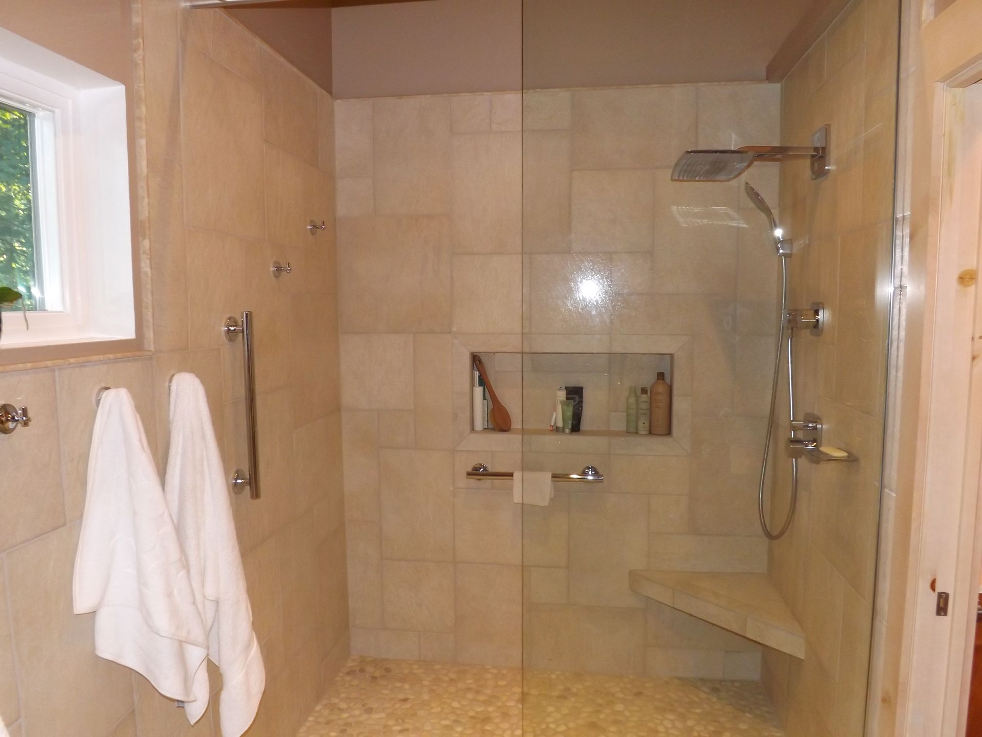 Walk-in shower with beige tile, glass shelf niche, wall-mounted showerhead, and two white towels on hooks