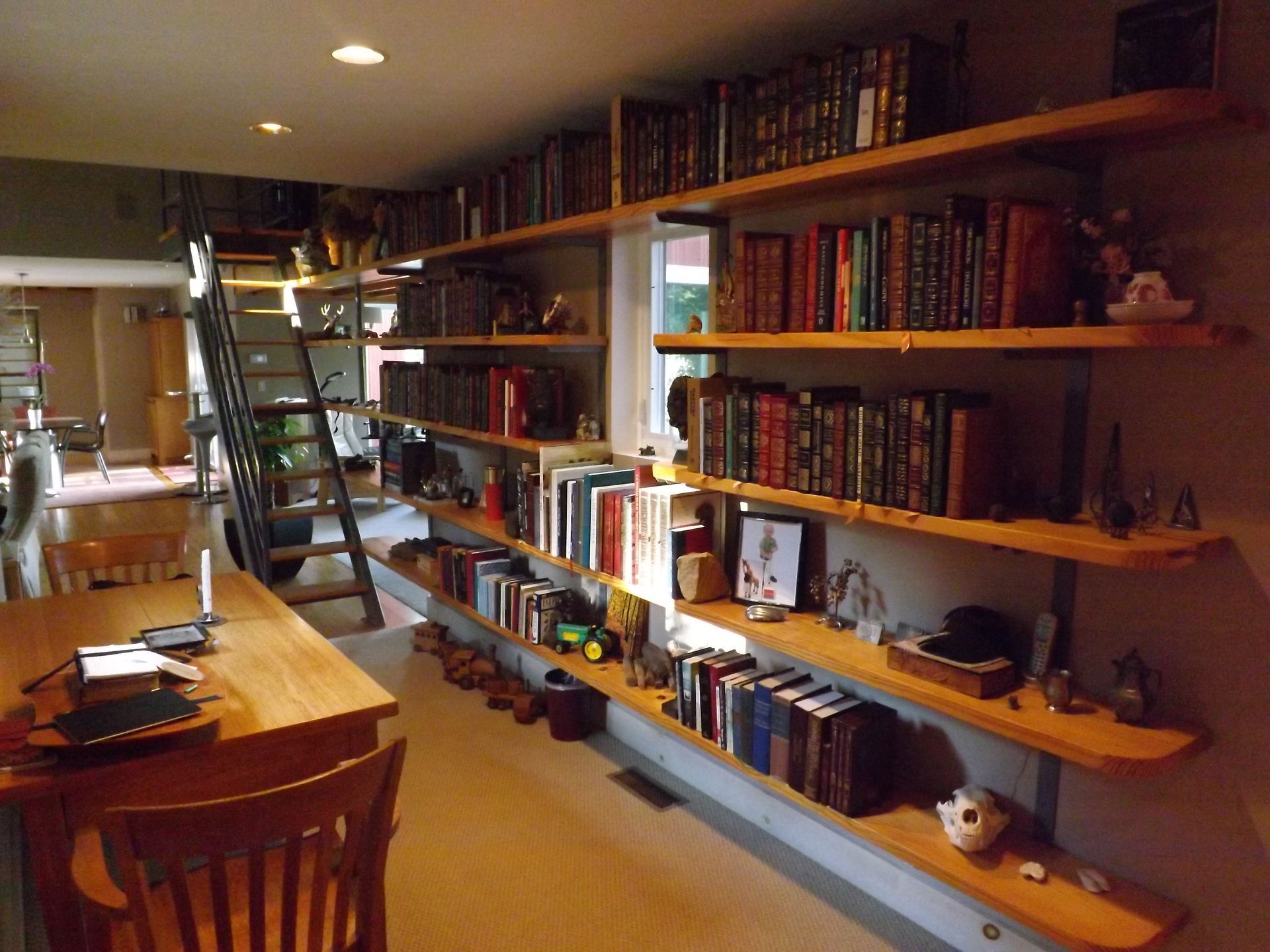 Cozy library room with wooden shelves full of books, a ladder, desk, and warm lighting.