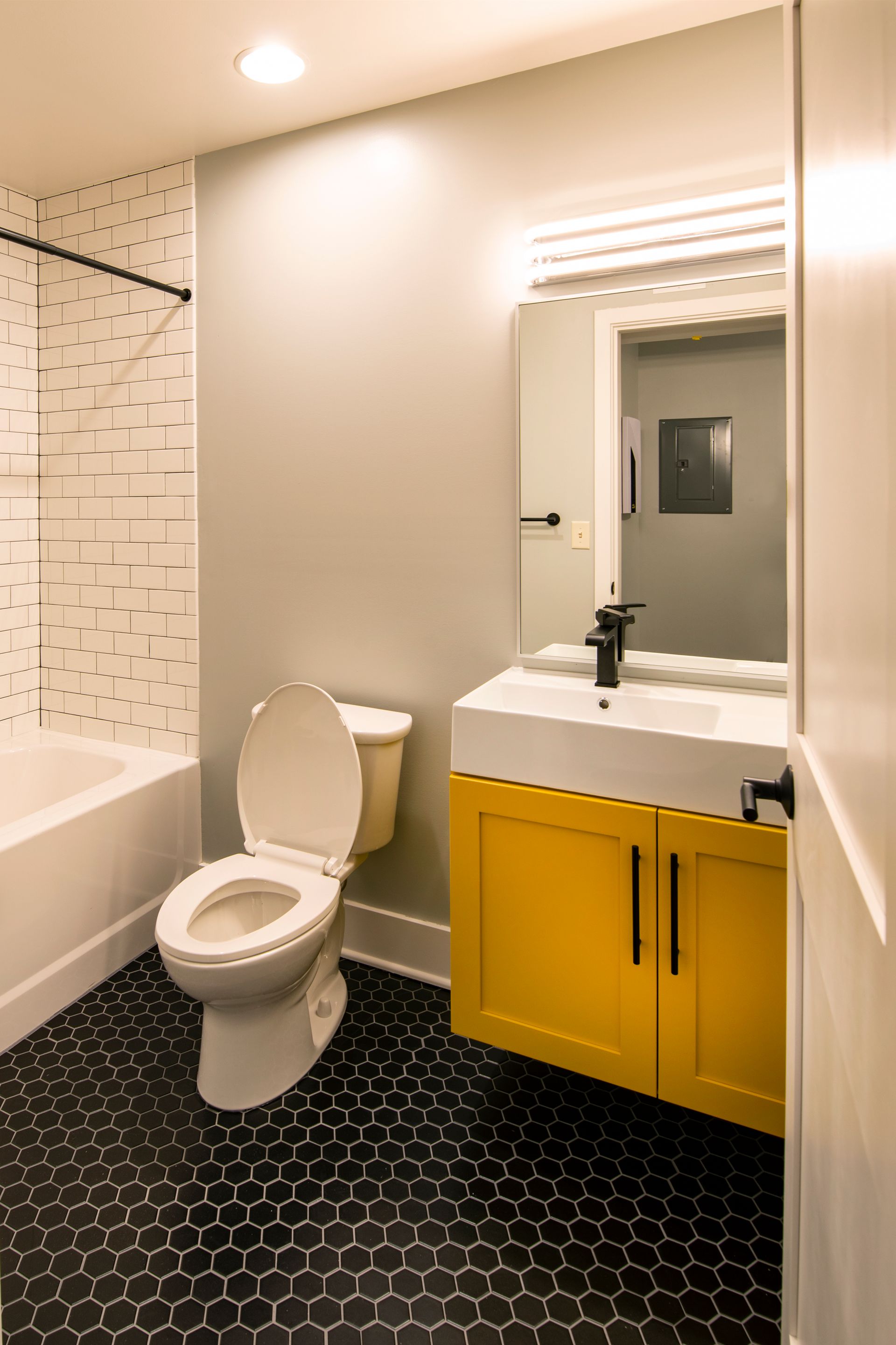 Modern bathroom with toilet, bathtub, white vanity, yellow cabinets, and black hex tile floor.