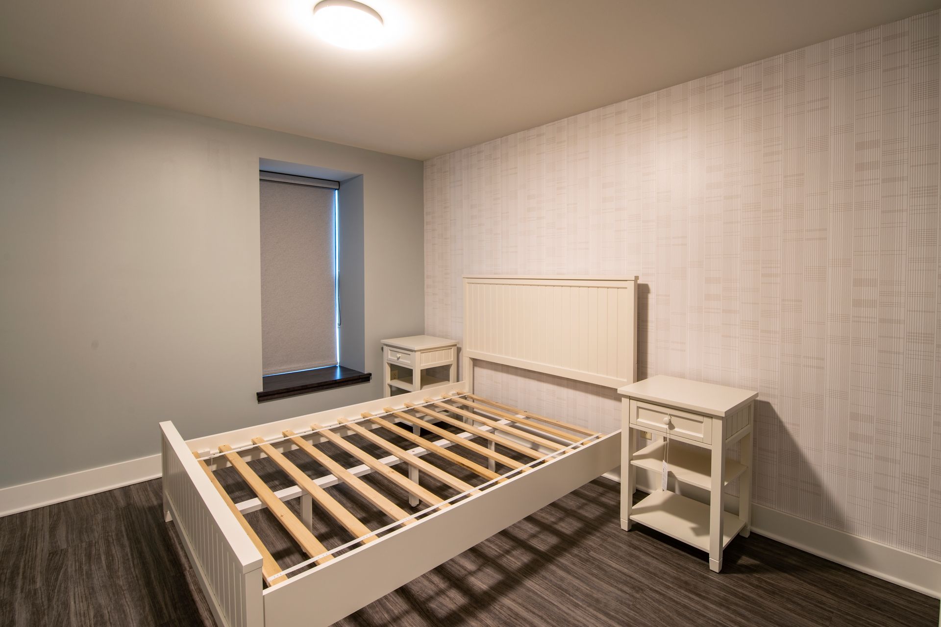 Empty bedroom with a bed frame, bedside table, and patterned wallpaper in soft gray tones.