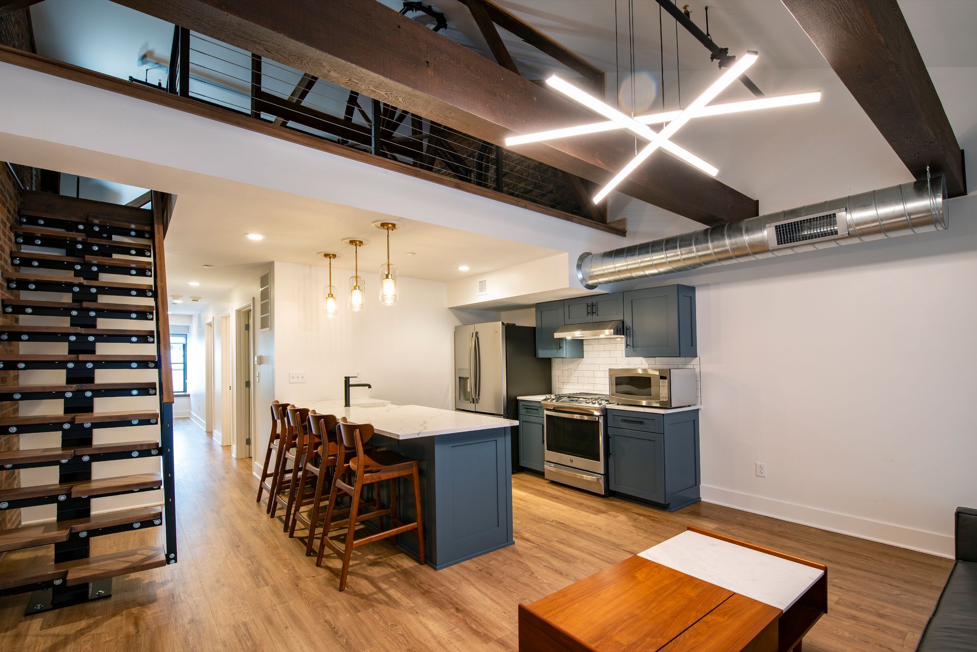 Modern loft kitchen with exposed beams, blue cabinets, bar stools, and a wooden staircase.