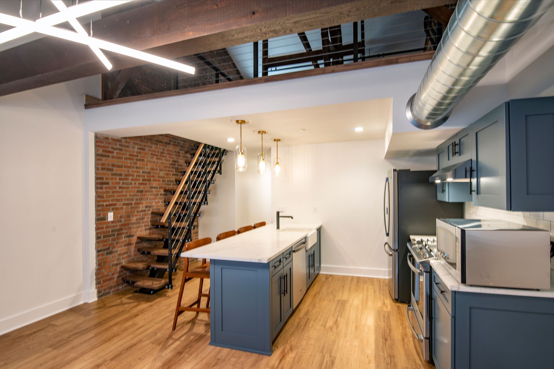 Modern kitchen with blue cabinets, wood floor, island, exposed brick wall, and staircase in a custom home.