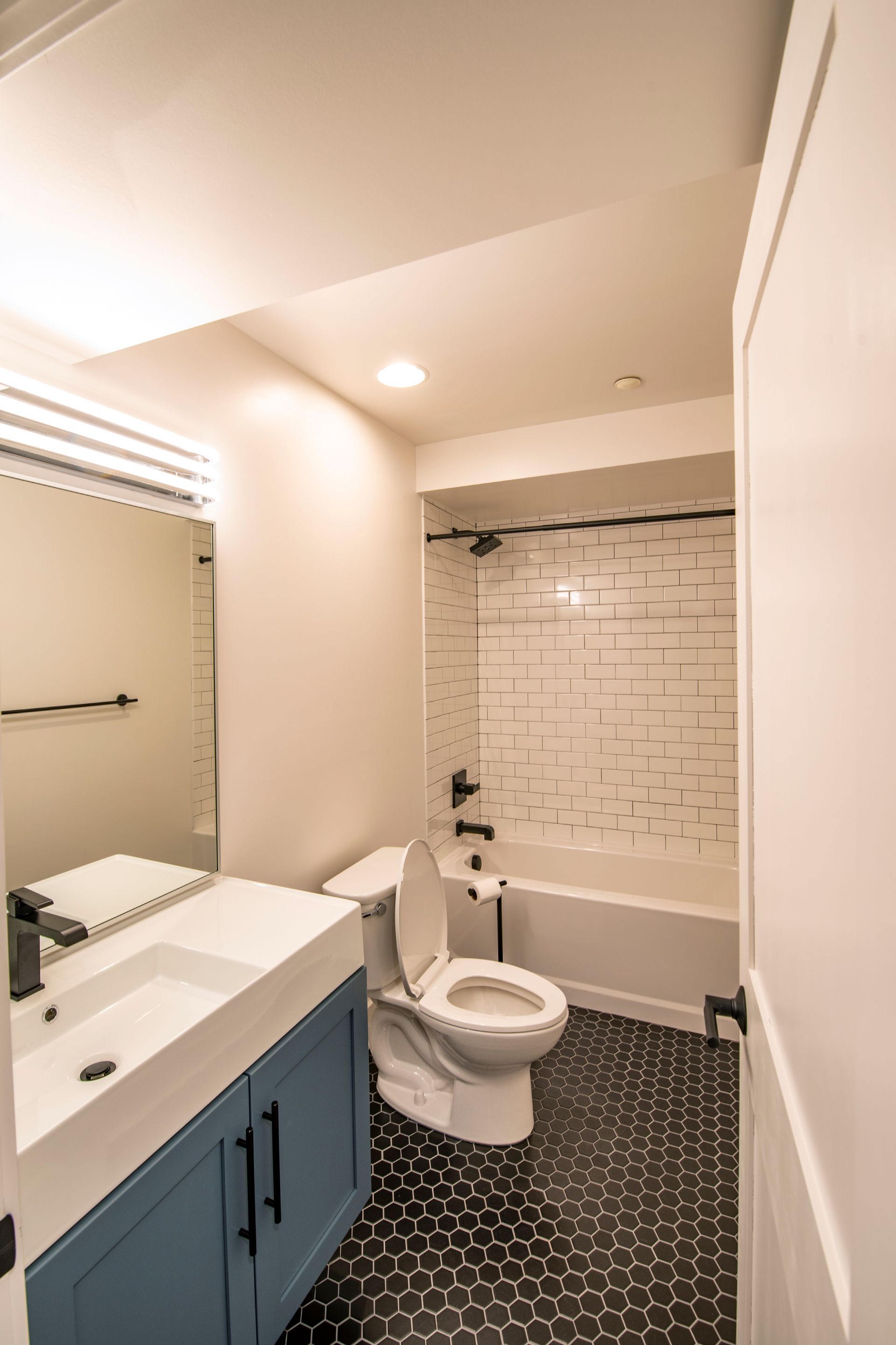 Small bathroom with blue vanity, white toilet, tiled shower, and patterned black floor.