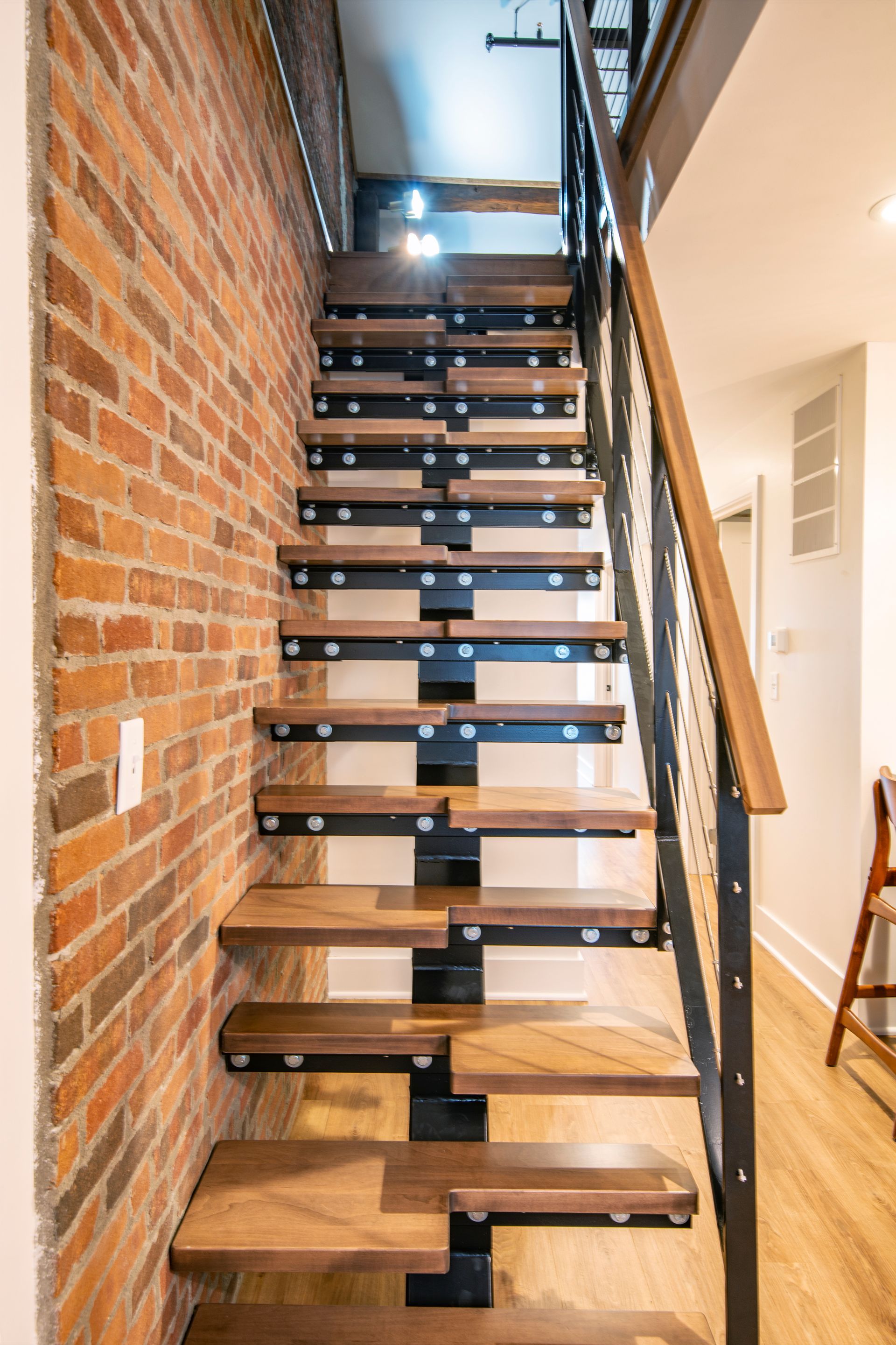 Narrow indoor staircase with wooden treads, black supports, and a brick wall beside it.