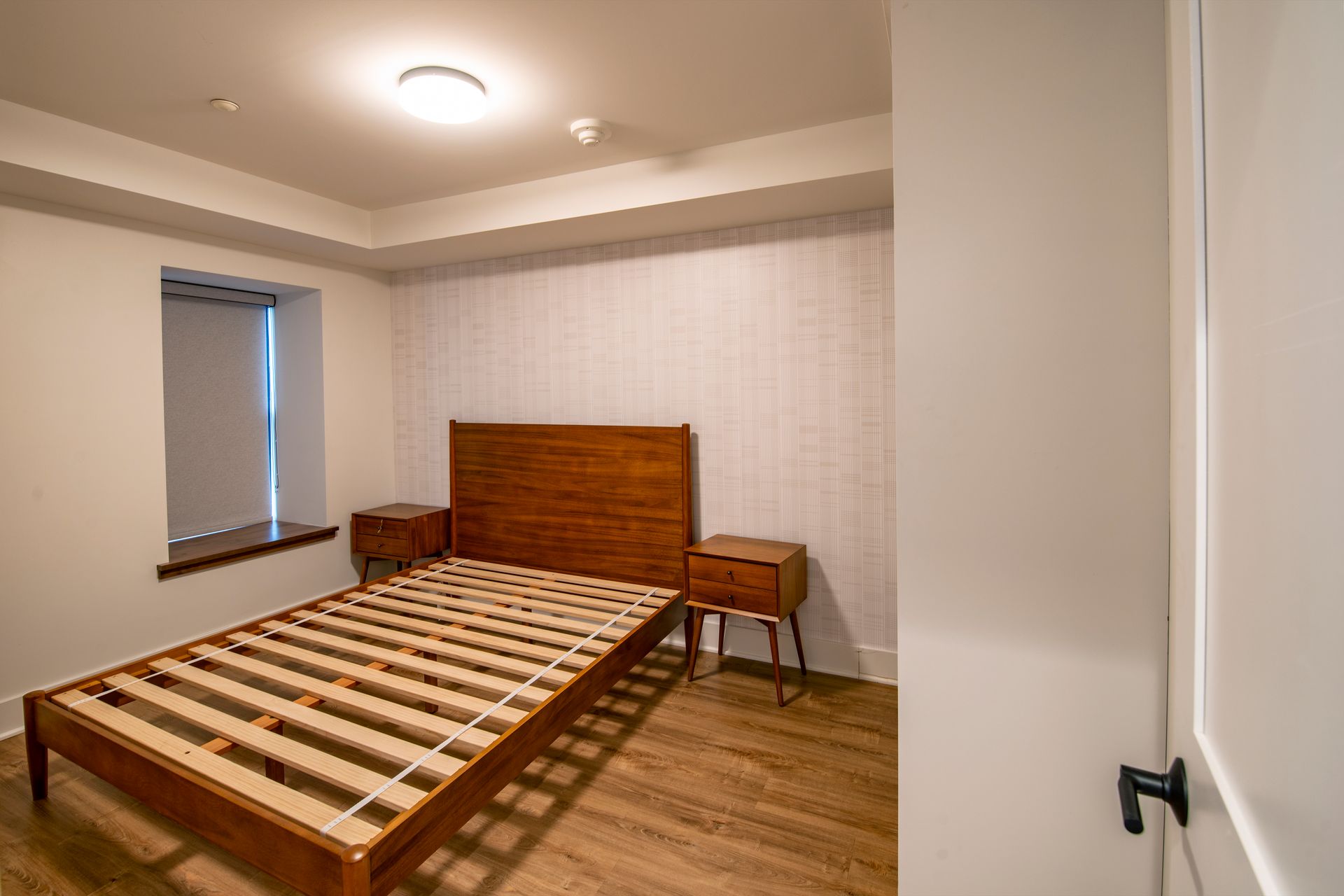 Simple bedroom with a bed frame, nightstand, small chair, and window blinds.