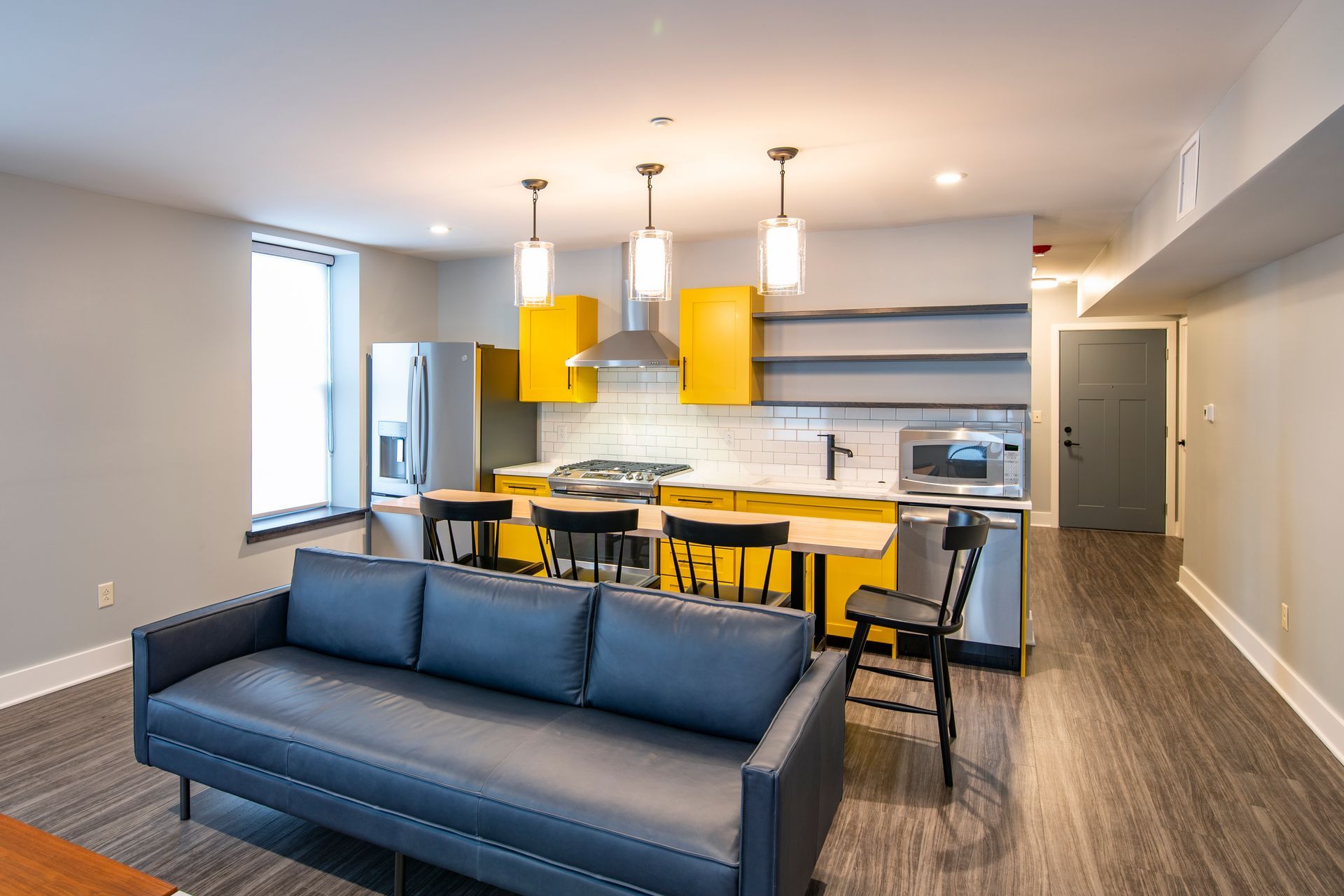 Modern apartment lounge with blue sofa, yellow chairs, and a kitchen area.