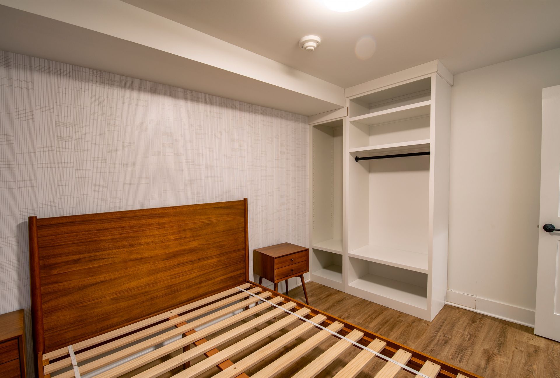 Empty bedroom with striped bed, wooden headboard, small nightstand, and open closet shelves.
