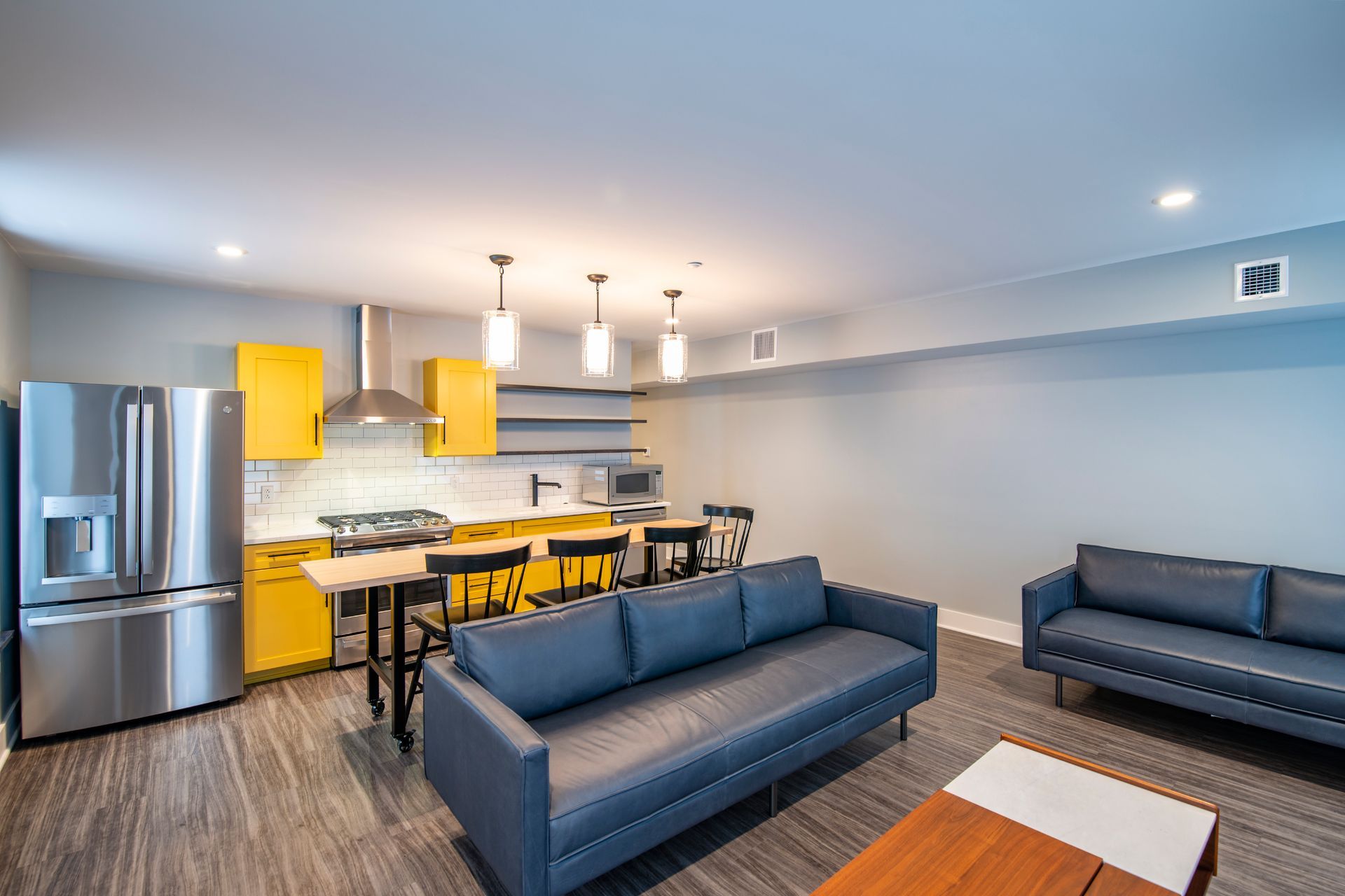 Modern open-plan kitchen and living room with stainless steel appliances, yellow chairs, and dark blue sofas.