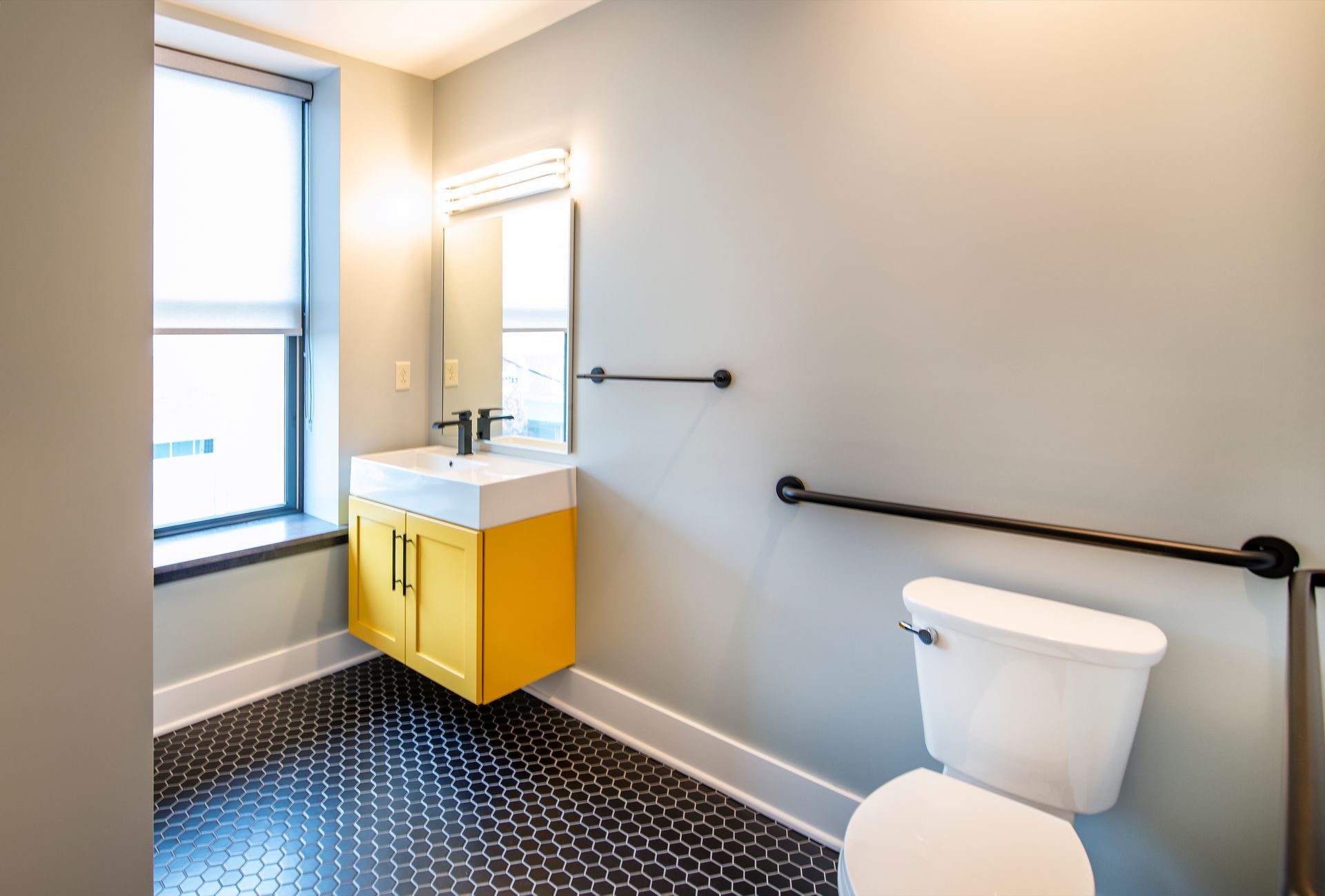 Accessible bathroom with yellow vanity, mirror, toilet, and black grab bars.