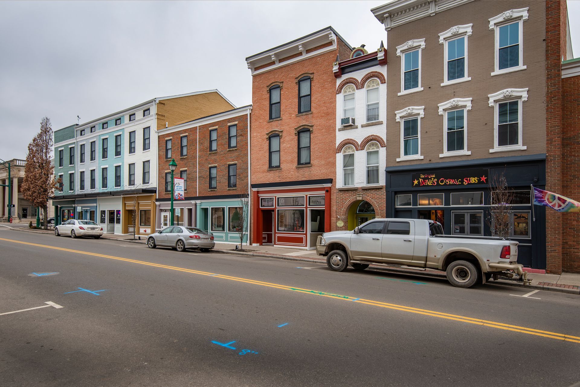 Small-town street with rowhouses, storefronts, parked cars, and a white pickup truck on the road.